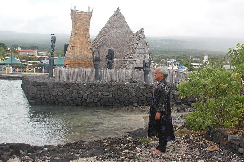 Alt text: A person stands on a rocky shoreline near the ocean in front of a straw-roofed Hawaiian temple that’s built on a black volcanic rock pier.
