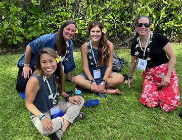 Four women sitting on grass smile at the camera. They all have yellow flowers in their hair and flower leis around their necks.
