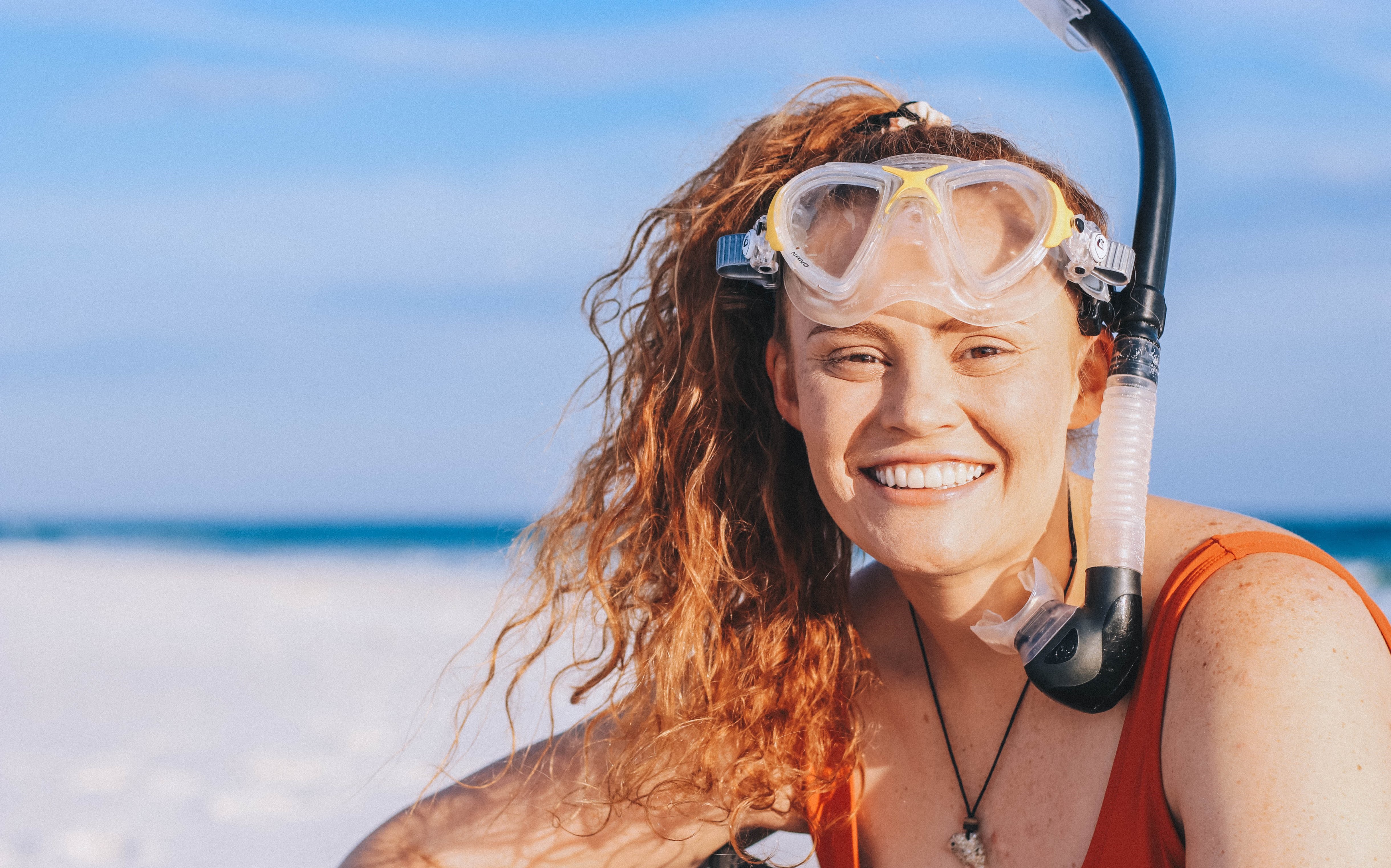 Woman with snorkel on her head smiling at the camera; beach in background.