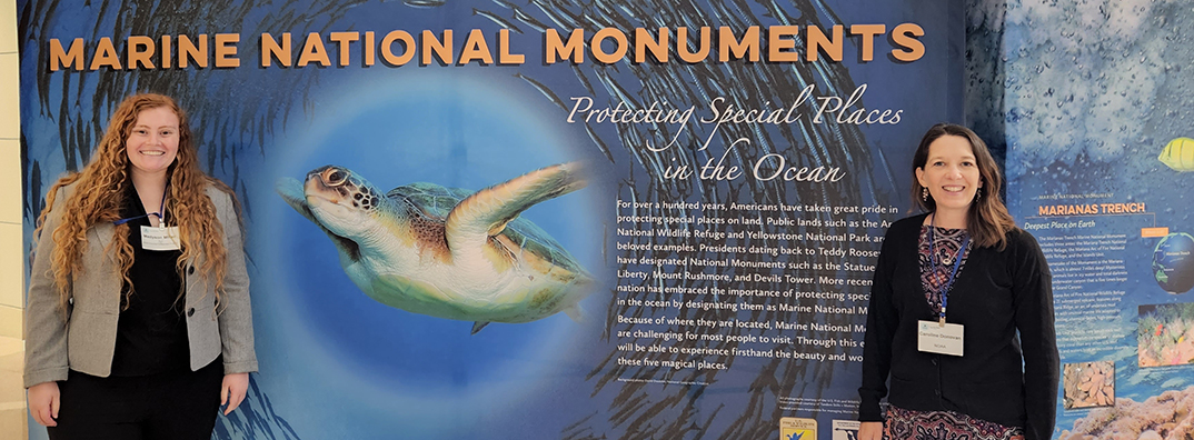 Two women standing on either side of a poster that reads 'Marine National Monuments' and shows a sea turtle in the center of a school of fish.