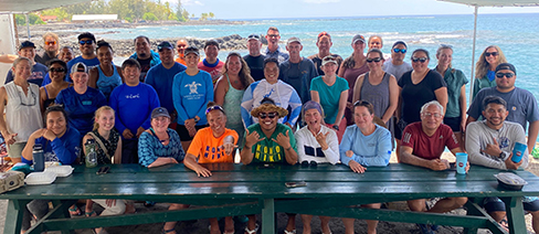 A large group of smiling people sitting at a table with the Hawaiian coastline behind them.