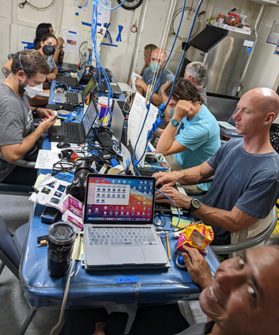 A group of people sitting at a table on a ship. They are looking at their laptop screens or smiling at the camera. Blue network lines hang from the ceiling and connect to each computer.