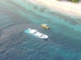  An aerial view of a deserted tropical coastline. A large white boat is half submerged in shallow water. A smaller yellow boat sits on the surface next to it.