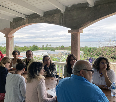 A group of people sit and talk on a covered porch overlooking green trees with the ocean in the background.